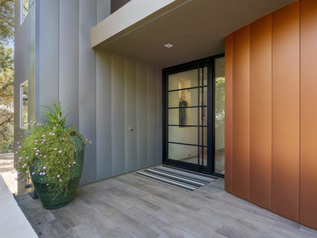modern entryway with glass pivot door, copper accent wall, vertical paneling, and large green planter with flowers