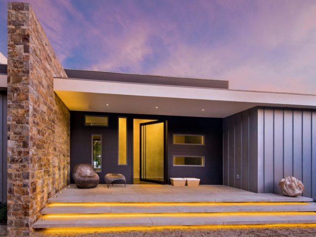 modern home entry with stone accent wall, glass pivot door, sleek design, and illuminated steps at sunset