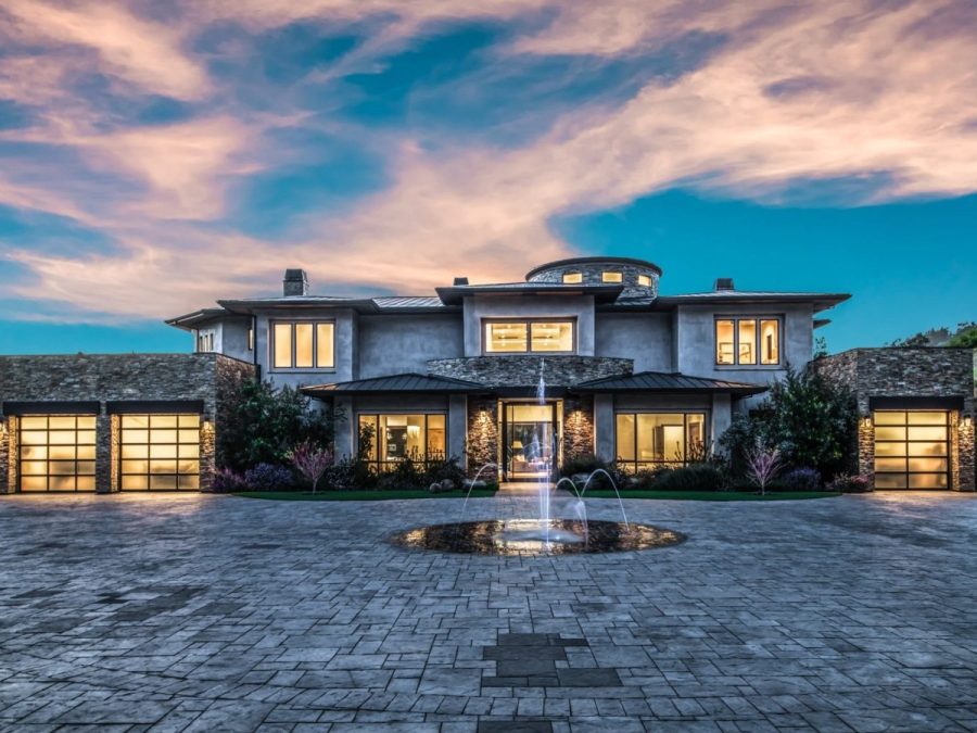 luxurious modern estate with stone and stucco siding, glass garage doors, circular driveway, and illuminated water fountain